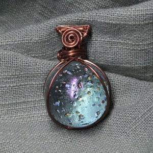 May include: A handcrafted pendant with a copper wire frame. The pendant features a blue and purple glass orb with a speckled design. The wirework includes a spiral at the top. The pendant is displayed on a gray fabric.