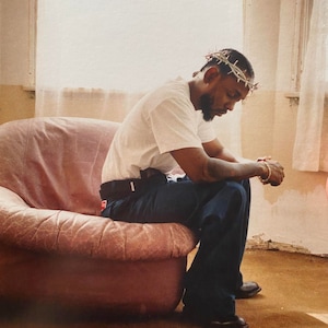 May include: A man wearing a white t-shirt and blue jeans sits on a pink armchair. He is wearing a crown of thorns on his head and has his hands clasped together.