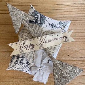 Puede incluir: Un adorno para tarta de aniversario hecho a mano con una flecha de purpurina plateada y una forma de copa de champán. El adorno presenta una pancarta que dice "Happy Anniversary" sobre una base en forma de estrella cubierta de tela con un lazo.