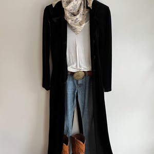 May include: A long black velvet duster coat worn over a white tank top and blue denim jeans. The outfit includes a tan patterned neck scarf, a brown leather belt with a large oval brass buckle, and a pair of brown leather cowboy boots.