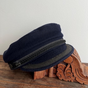 May include: A navy blue wool cap with a black braided band and a black leather brim. The cap is resting on a carved wooden decorative piece.