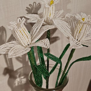 May include: A glass vase holds three beaded white flowers with yellow centers and green stems and leaves. The flowers are made of small white beads, and the stems and leaves are made of green beads. The vase is clear and sits against a neutral background.