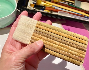 Puede incluir: Un conjunto de 10 pinceles de caligrafía de bambú con cerdas blancas. Los pinceles están agrupados y se sostienen en una mano.