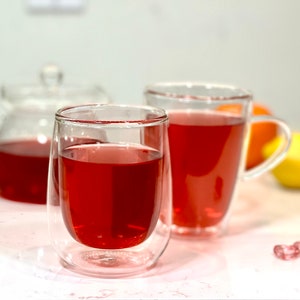 May include: Two clear glass mugs with double walls filled with red tea. The mugs are on a white surface with a few red heart-shaped decorations.