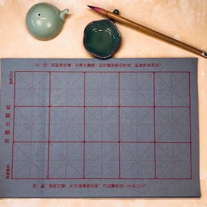 May include: A gray calligraphy practice sheet with red grid lines. The sheet has Chinese characters printed on the top and bottom. The sheet is surrounded by a brush pen, a small green inkwell, and a small green water pot.