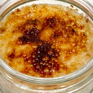 Puede incluir: Un tarro de vidrio lleno de un postre blanco cremoso cubierto con una capa de azúcar caramelizado. El azúcar tiene una textura burbujeante y un color marrón intenso.