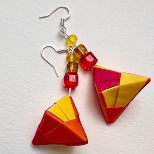 Op de afbeelding: Een paar origami-oorbellen van rood, geel en oranje papier. De oorbellen zijn bevestigd aan zilveren haakjes met een cluster van rode, gele en amberkleurige kralen.