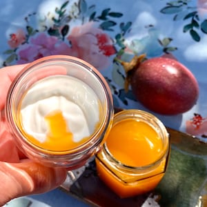 May include: A glass jar filled with a bright orange liquid, next to a glass cup with a white yogurt base and a layer of orange liquid on top. The jar and cup are on a brown and green tray.