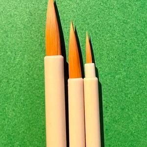 May include: Three paintbrushes with white handles and natural bristles of varying sizes. The brushes are arranged in order of size, with the largest brush on the left and the smallest brush on the right. The brushes are set against a green background.