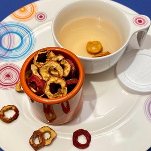 May include: A white teacup with a handle and a small orange pot filled with dried fruit rings sit on a white plate with colorful concentric circle designs. The cup contains a light-colored liquid and a few pieces of fruit.