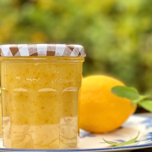May include: A jar of orange-yellow marmalade with a checkered lid sits on a blue and white plate next to a whole lemon with green leaves. A smaller jar of marmalade is in the background.