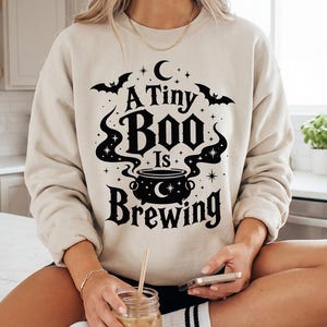 May include: A beige crewneck sweatshirt featuring a black graphic print of a cauldron with swirling smoke, bats, stars, and a crescent moon. The text reads A Tiny Boo Is Brewing in a decorative font. The sweatshirt has long sleeves and ribbed cuffs.