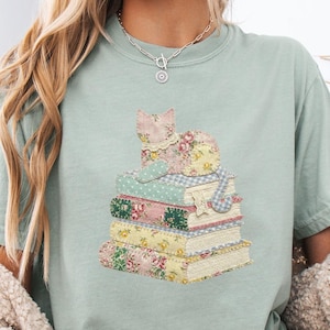 May include: A light green t-shirt featuring a stack of books with a floral pattern and a cat design. The cat and books have a patchwork design with pink, yellow, and blue floral patterns. The shirt has a relaxed fit.