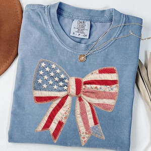 May include: A light blue Comfort Colors t-shirt featuring a large bow design. The bow is made of fabric patches in red, white, and blue, with a star pattern on the left side. A gold necklace with a pendant is draped across the shirt.