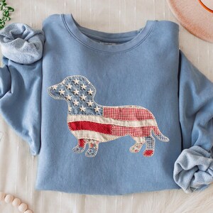 May include: A light blue sweatshirt featuring a patriotic dachshund design. The dog is crafted from fabric patches, incorporating the American flag's stars and stripes. The sleeves are cuffed, and the sweatshirt is displayed on a white surface.