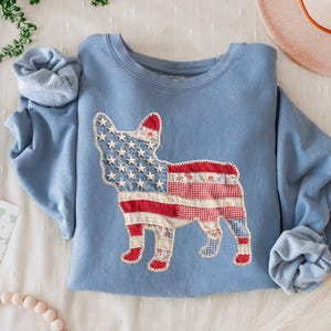 May include: A light blue sweatshirt featuring a patchwork design of a dog in the style of the American flag. The dog's body is made of red, white, and blue patterned fabrics. The text "BLUE JEAN" is at the bottom.