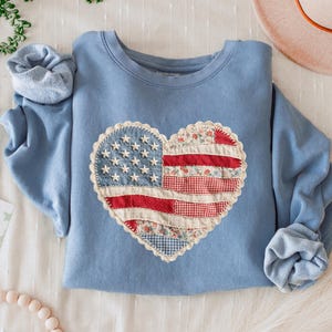 May include: A light blue sweatshirt featuring a heart-shaped American flag design. The flag is made of various fabric patterns, including stars, stripes, and floral prints. The sleeves are cuffed, and the text "BLUE JEAN" is at the bottom.