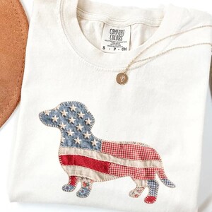 Ivory Comfort Colors patriotic dachshund patchwork t-shirt for women, featuring a large quilted wiener dog graphic with embroidered white stars, denim blue, red gingham, cream floral, and striped fabric — 4th of July dachshund mom gift, American flag wiener dog tee, cute dog mom patriotic shirt, patchwork dachshund Comfort Colors tee, dachshund lover gift idea.