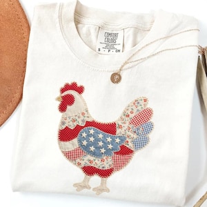 May include: Cream-colored Comfort Colors t-shirt featuring a patchwork chicken design with red, white, and blue patterns. A gold necklace with a pendant rests on the shirt. The shirt is folded, and a brown hat is partially visible.