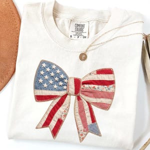 Ivory Comfort Colors patriotic bow patchwork t-shirt for women, featuring a large quilted bow graphic with embroidered white stars, red gingham, cream floral, and striped fabric — coquette 4th of July gift idea, American flag bow tee, cute patriotic shirt women, patchwork bow Comfort Colors shirt.