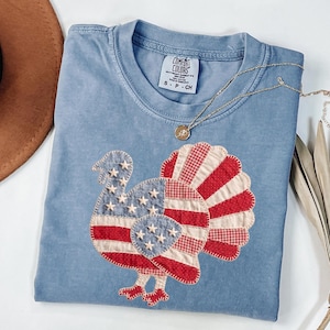 May include: A blue short-sleeved t-shirt featuring a large turkey applique on the front. The turkey is designed with red and white stripes and a blue section with white stars, mimicking an American flag pattern. The shirt has a Comfort Colors label.