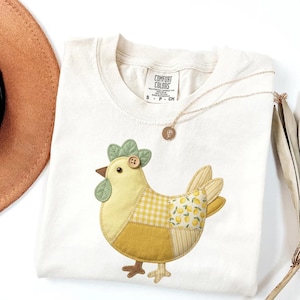 May include: Cream-colored Comfort Colors t-shirt featuring a patchwork chicken design in shades of yellow and green. The chicken has a button eye and a green leafy comb. A gold necklace with a pendant rests on the shirt. The shirt is folded.