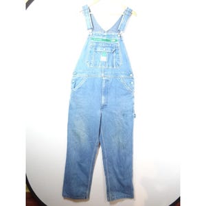 May include: Light blue denim overalls featuring a bib pocket with a green zipper closure, adjustable shoulder straps with metal buckles, side buttons, and two front pockets. The garment has a classic straight-leg cut with visible stitching.