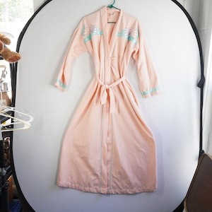 May include: A peach-colored robe with a matching belt, hanging on a green hanger. The robe features light blue and floral embroidery on the shoulders and cuffs. Vertical light blue stripes run down the front of the robe.