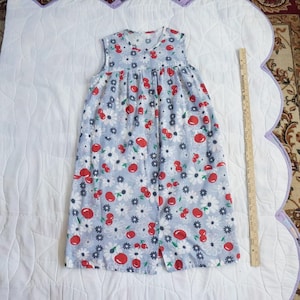 May include: A sleeveless, light blue dress with a cherry and daisy pattern. The dress has a V-neck and a button-down front. The fabric is covered in red cherries, white daisies, and small blue flowers. The dress is laid out on a white quilted surface.