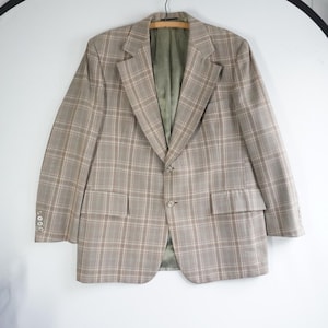 May include: A tan plaid blazer with a two-button closure and two front pockets. The blazer has a notched lapel and three buttons on each cuff. The interior lining is a solid olive green color. The blazer is hanging on a wooden hanger.