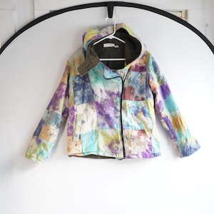 May include: A multi-colored, patchwork-style jacket with a hood, hanging on a white hanger. The jacket features a zipper closure and a mix of colors including purple, blue, yellow, and green. The jacket is made of a soft material.