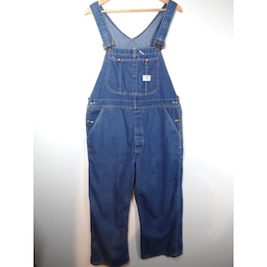 May include: Medium-wash blue denim overalls featuring a bib pocket with a small white rectangular brand label, adjustable shoulder straps with metal buckles, side button closures, and a classic straight-leg cut with visible copper-toned rivets.