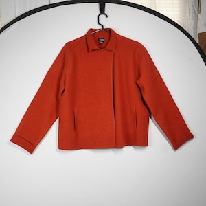 May include: An orange-red jacket with a collar and two front pockets. The sleeves are long with folded cuffs. The jacket is hanging on a black hanger against a white background.