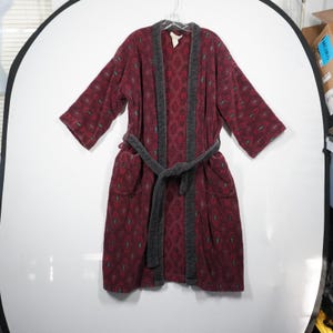 May include: A burgundy robe with a geometric pattern and gray trim. The robe has a matching belt and pockets. The robe is hanging on a hanger against a white background. The robe is made of a soft, plush material.