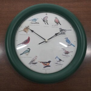 May include: A round wall clock with a dark green frame and a white face featuring illustrations of twelve different bird species arranged around the dial. Each bird is labeled with its common name, and the clock has three black hands.