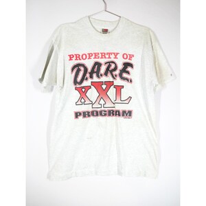 May include: A light gray short-sleeve t-shirt featuring the red and black text PROPERTY OF D.A.R.E. XXL PROGRAM printed on the front. The shirt has a crew neckline and is displayed on a metal hanger against a plain white background.