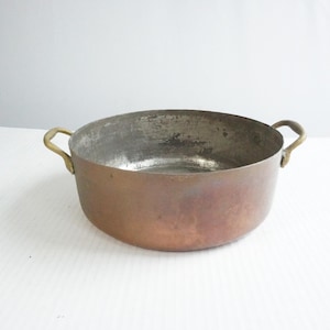 May include: A copper-colored, round cooking pot with two brass handles. The pot has a tarnished interior and a slightly distressed exterior, suggesting age and use. The pot is likely vintage or antique.