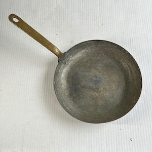 May include: A vintage, round frying pan with a long, brass handle. The pan's cooking surface is a dark, aged metal, showing signs of use. The handle has a hole for hanging. This cookware is ideal for rustic kitchen decor.