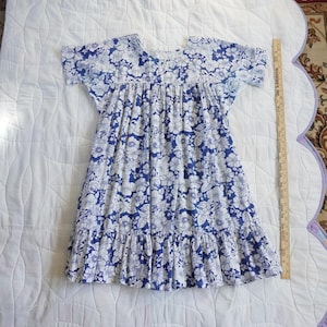 May include: A short-sleeved dress with a blue floral pattern on a white background. The dress has a ruffled hem and a gathered bodice. A wooden ruler is placed next to the dress.