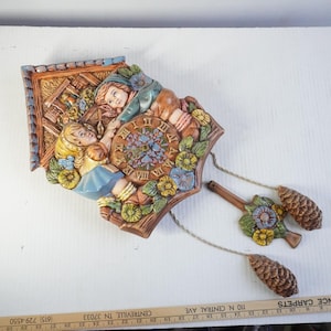 May include: A vintage cuckoo clock with a detailed relief of children and flowers. The clock face features Roman numerals and a floral design. The pendulum has a floral design and pinecone weights. The clock is brown, blue, yellow, and green.