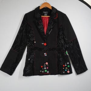 May include: Black blazer with floral pattern accents and red stitching. The blazer features a red interior lining, decorative buttons, and a unique design. The blazer is hanging on a wooden hanger.