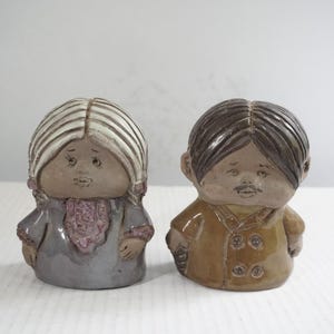 May include: Two ceramic figurines, a man and a woman, stand side-by-side. The woman has white hair and wears a gray dress with a purple scarf. The man has brown hair and a mustache, wearing a tan coat.