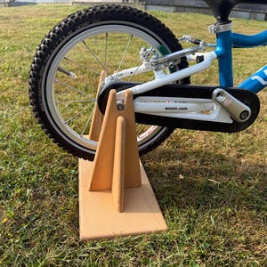 Könnte beinhalten: Ein blau-weißes Kinderfahrrad mit schwarzen Reifen steht auf einem Holzständer. Das Fahrrad hat einen weißen Rahmen, einen schwarzen Kettenschutz und die Marke "woom" ist auf dem Rahmen sichtbar. Der Ständer ist aus hellem Holz.