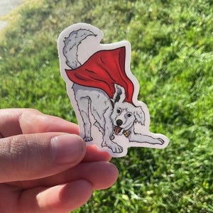May include: A sticker of a white dog wearing a red cape, with its tongue out and a gold collar. The dog is in a playful pose, with its front paws extended. The sticker has a white border and is set against a green background.