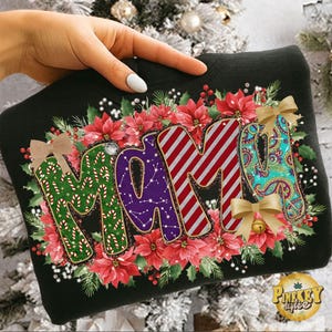 Könnte beinhalten: Schwarzes Sweatshirt mit dem Wort "MAMA" in einem festlichen Design. Die Buchstaben sind mit Zuckerstangen, Streifen, Sternbildern und Paisley-Mustern verziert, umgeben von roten Weihnachtssternen und Grün. Goldene Schleifen und eine kleine Glocke ergänzen das festliche Thema.