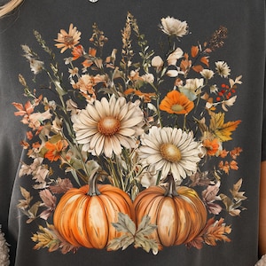 Puede incluir: Camiseta gris oscuro con un diseño floral y de calabaza. El diseño incluye margaritas blancas, amapolas naranjas y dos calabazas naranjas. La camiseta tiene un aspecto vintage y desgastado.