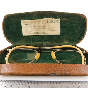 May include: Vintage metal-framed eyeglasses with yellow temple covers and nose pads, resting inside a brown textured hard case with a green velvet interior. A white label inside the case displays the text 108925, date 2-25-48, and address details.