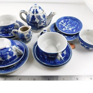 May include: A miniature tea set featuring a white teapot, creamer, and four cups with saucers, all decorated with a blue willow pattern. A metal ruler at the bottom shows the set spans approximately 10 inches in total width across the arrangement.