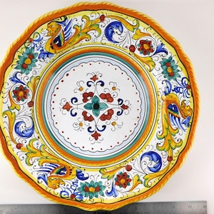 May include: A round ceramic plate with a scalloped edge, featuring a white center with a red and teal floral motif. The border displays yellow, blue, and green scrollwork and mythical creatures. A metal ruler shows a diameter of 12 inches.