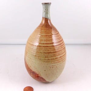 May include: A ceramic vase with a bulbous body and a narrow neck, featuring a glossy glaze in shades of tan, cream, and reddish-brown with horizontal ridges. A copper-colored coin is placed at the base for scale reference.